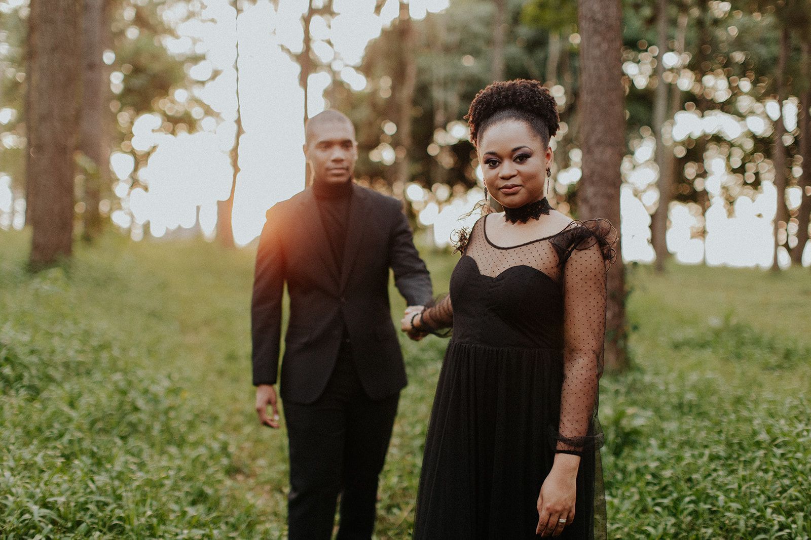 A man in a suit and a woman in a black dress are holding hands in the woods.