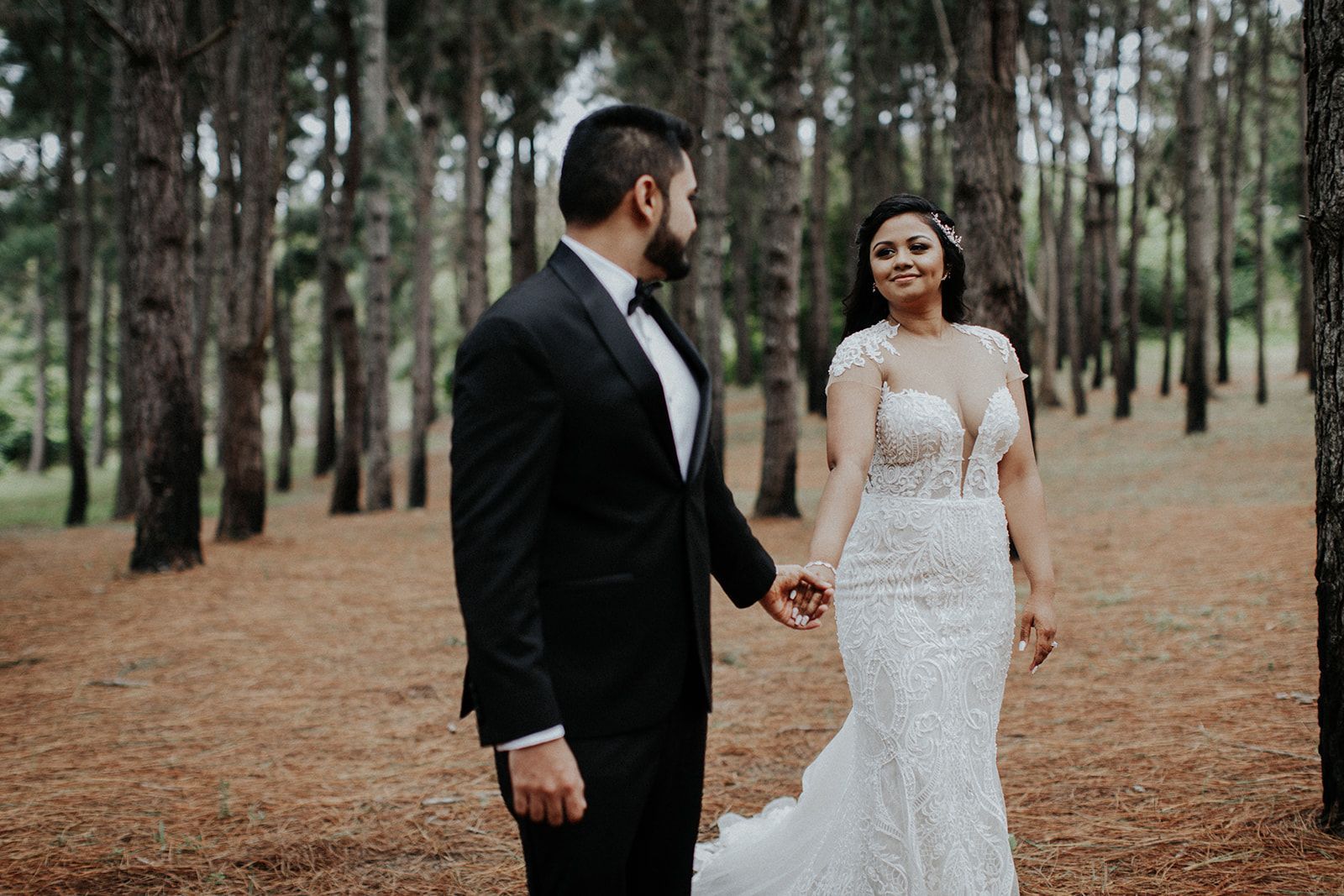 A bride and groom are walking through a forest holding hands.