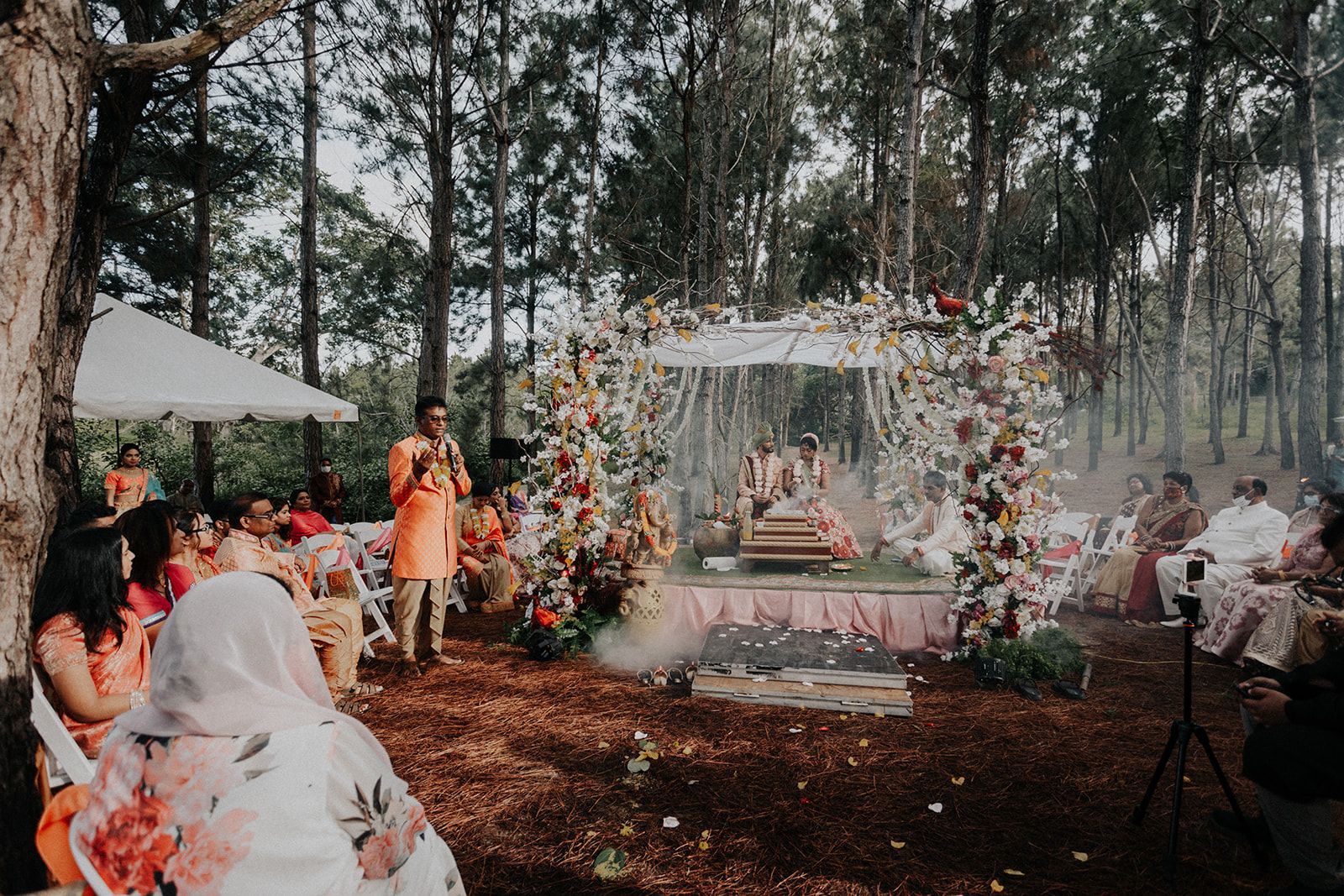 A group of people are sitting under a tent in the woods at a wedding ceremony.
