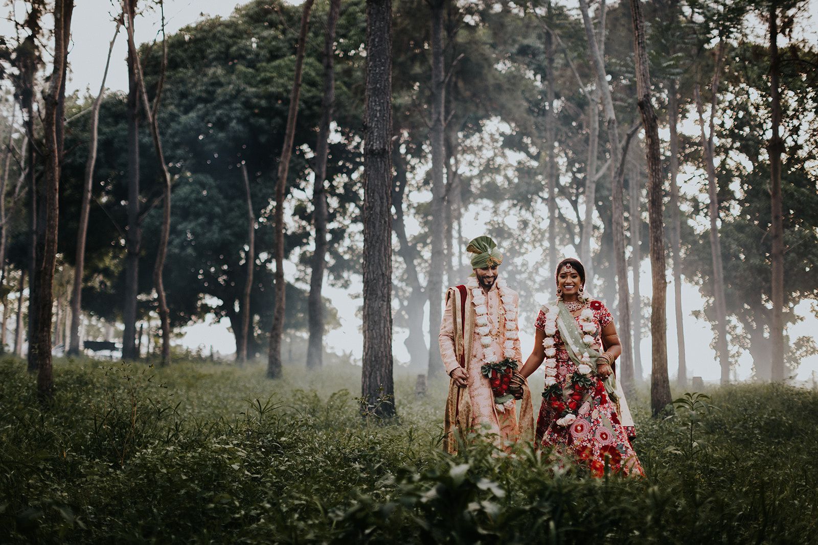 A bride and groom are walking through a forest holding hands.
