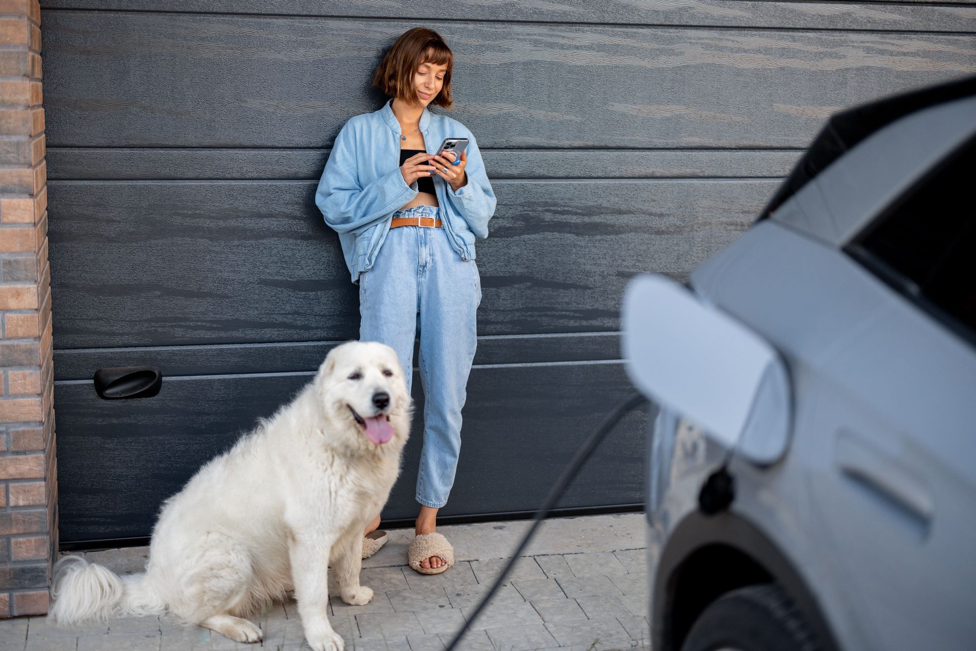 A woman stands leaning against a gray garage door, looking at her smartphone, while a large white do