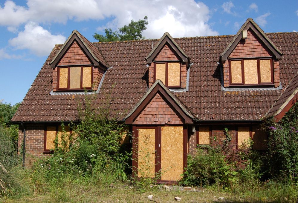 Boarded-up brick house with overgrown bushes, three roof dormers, and blue sky.