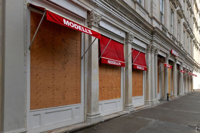 Boarded-up Modell's storefronts with red awnings along a city street.