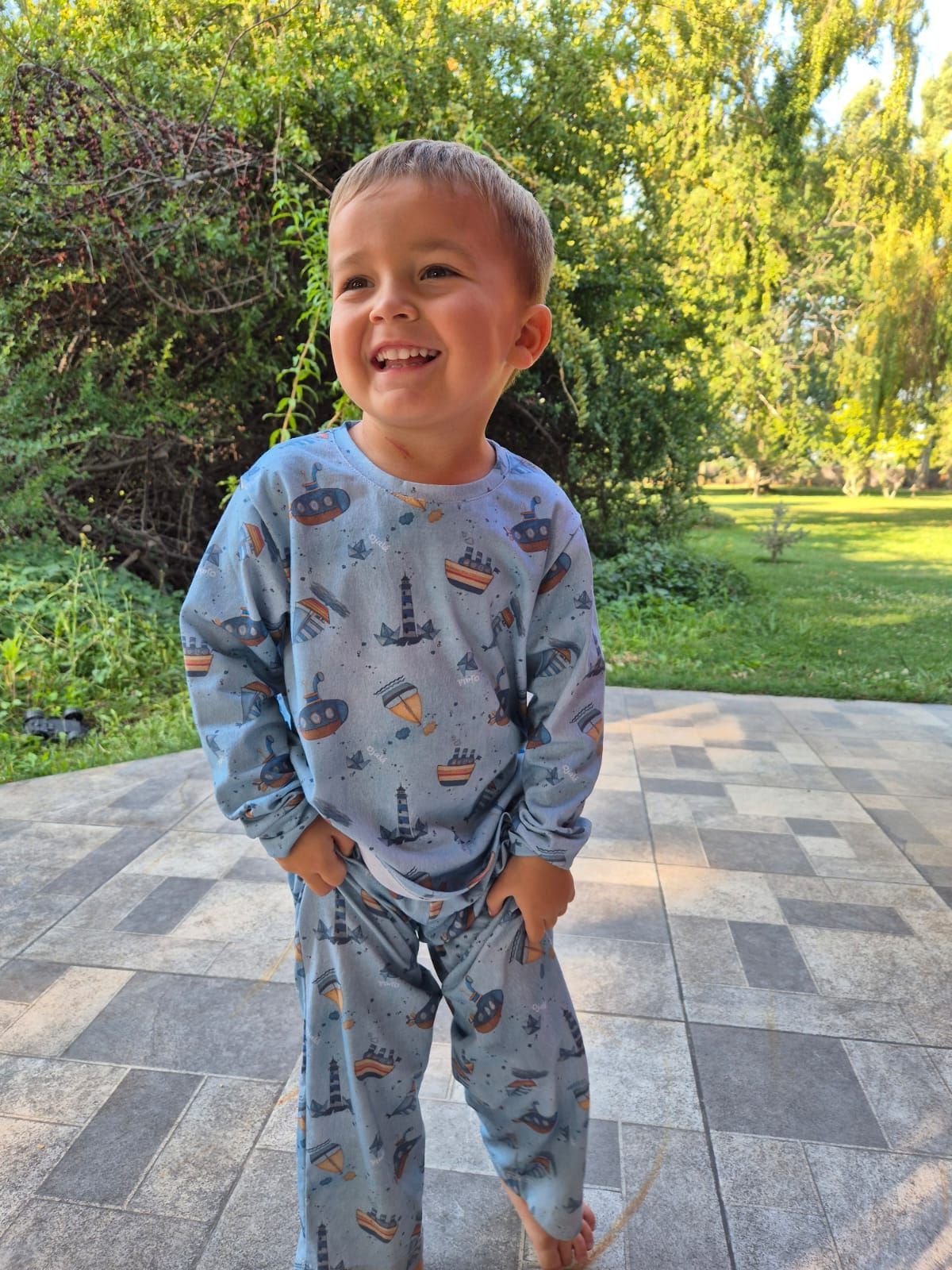 Niño pequeño sonriendo, vistiendo un pijama azul de temática náutica, de pie afuera en un patio.
