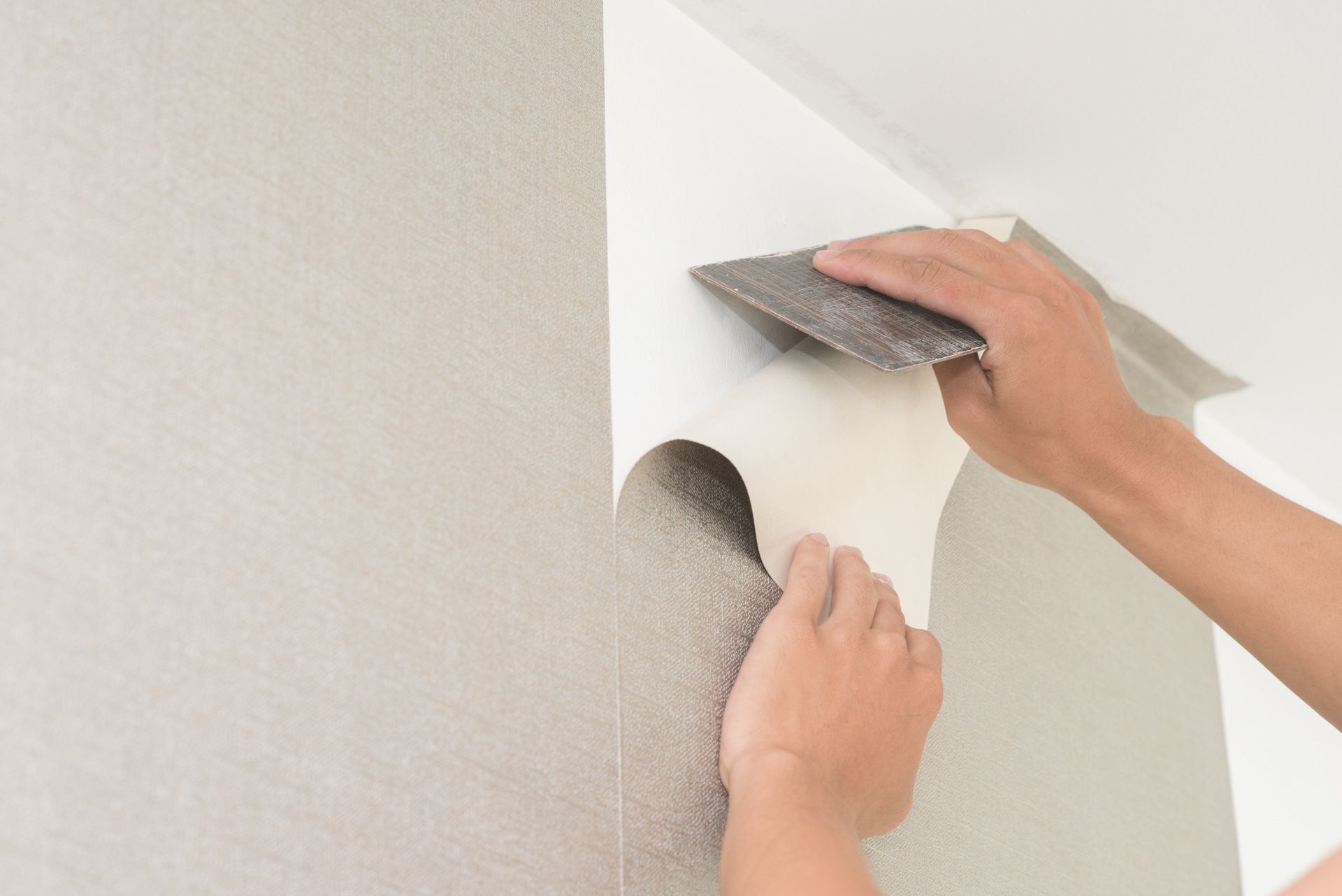 A person is peeling wallpaper from a wall with a spatula.