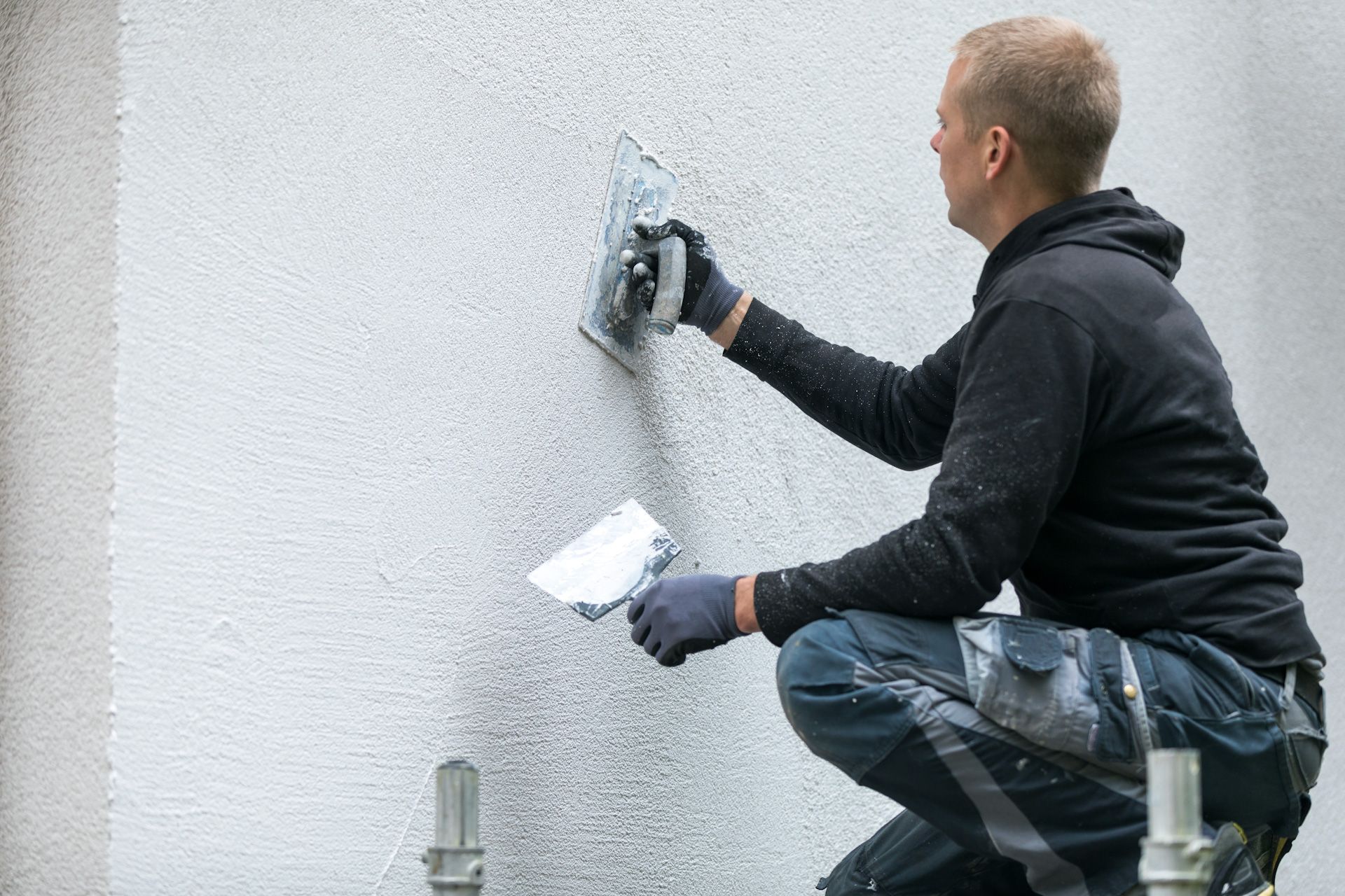 A man is kneeling down on a scaffolding while using a spatula on a wall.