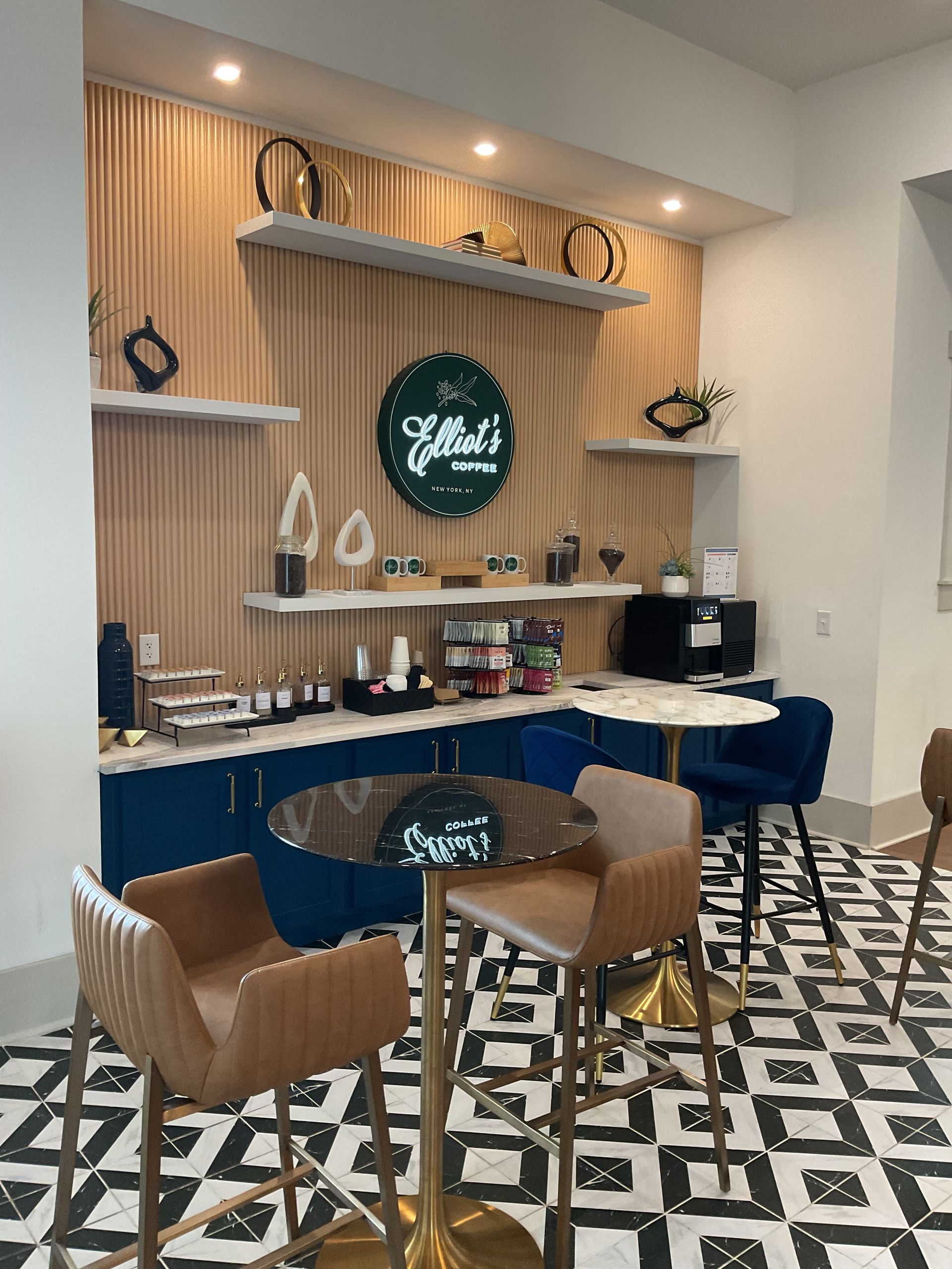Coffee bar interior with blue cabinets, tan bar stools, and decorative shelves on a wood-paneled wall.