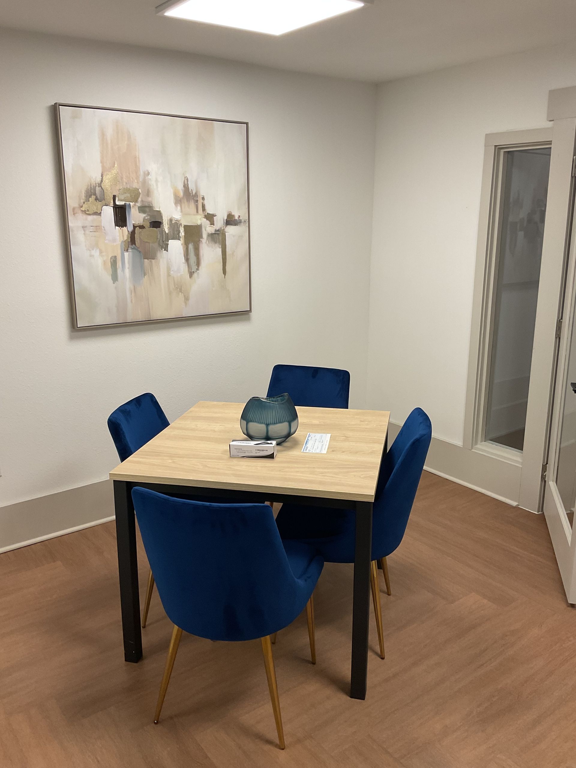 A small dining area with a wood table, four blue velvet chairs, and abstract art.