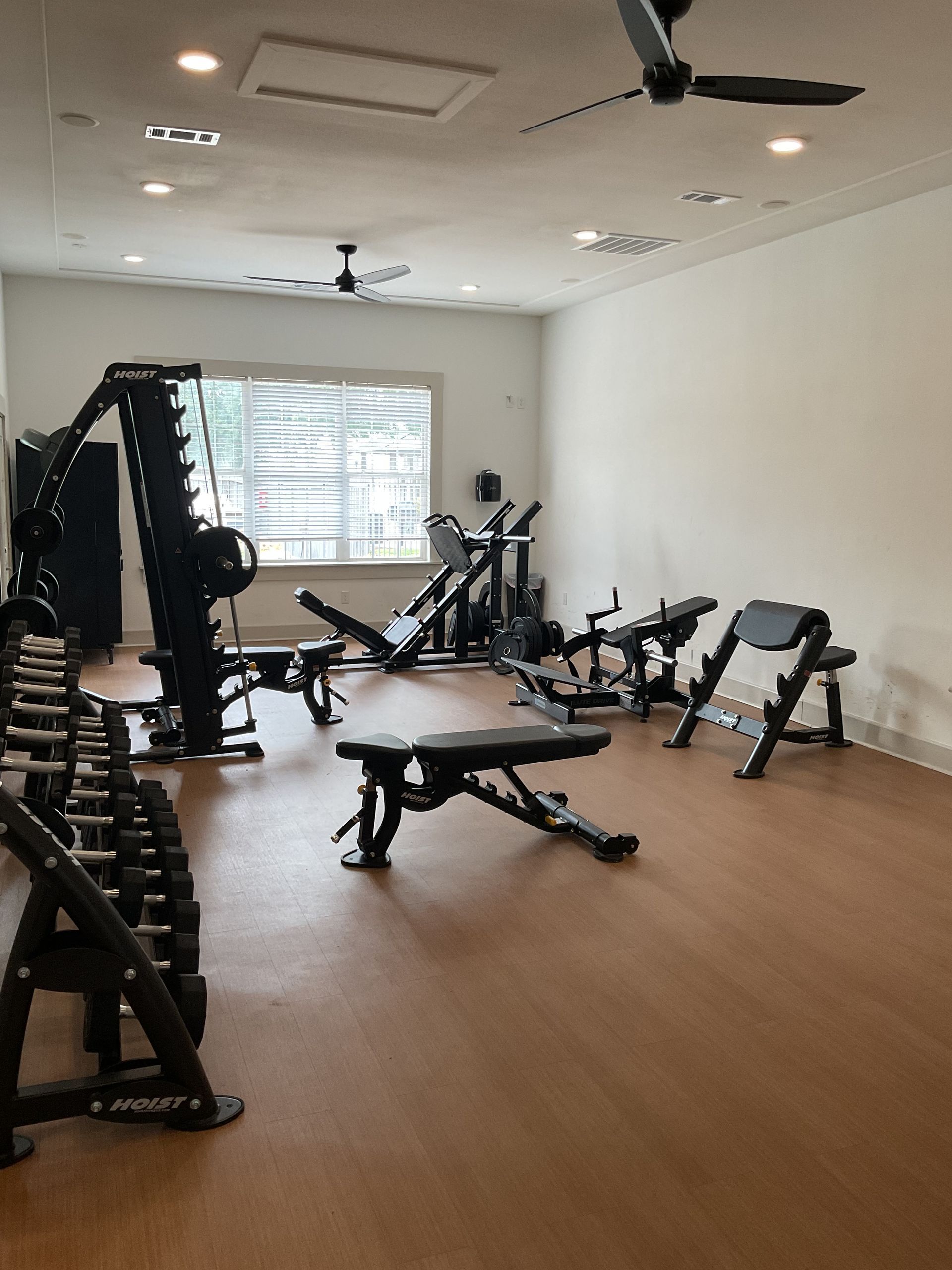 Gym interior with weight machines, dumbbells, benches, and a window. Brown floor, white walls.