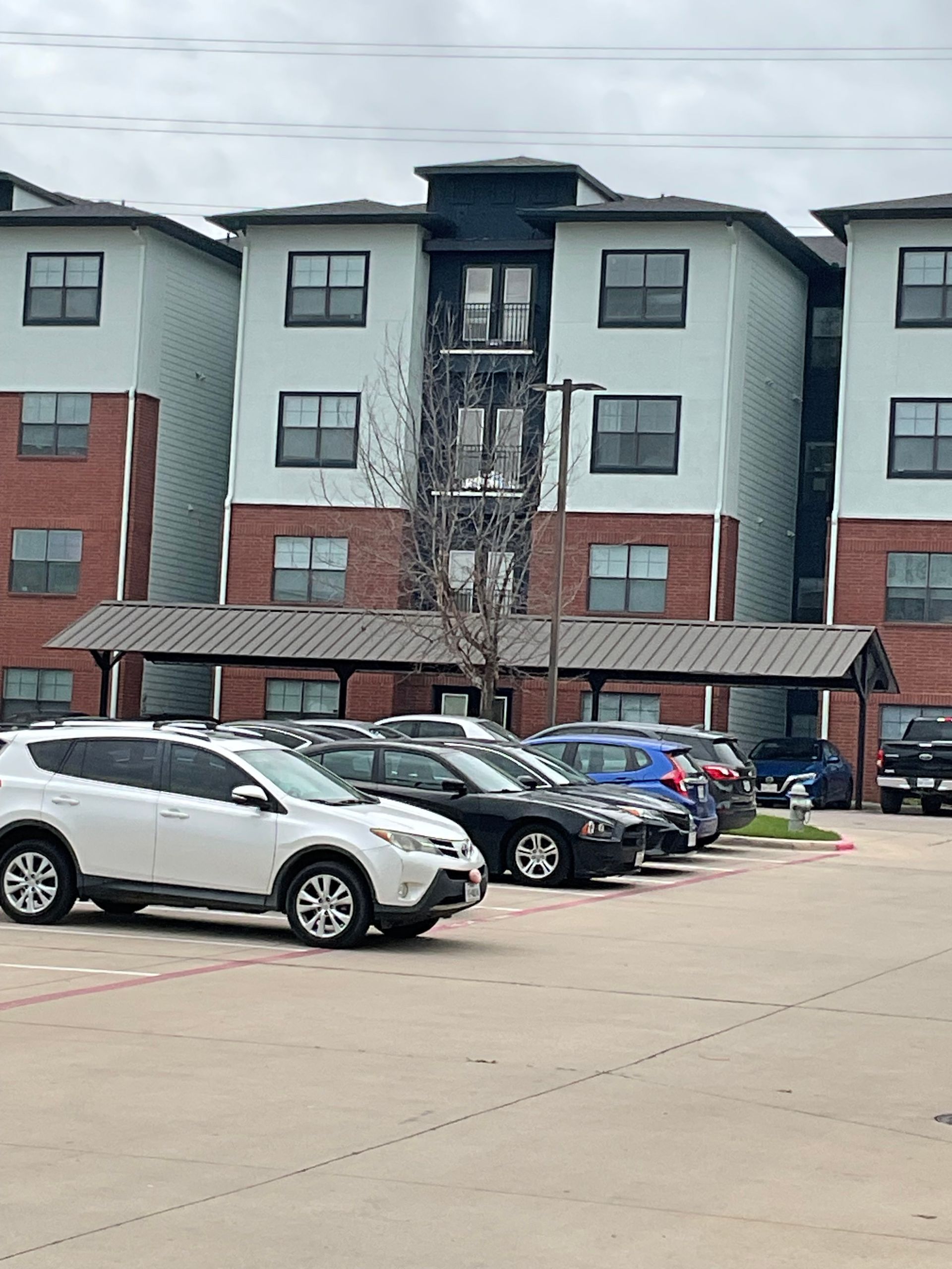 Apartment building with cars parked in front. Brick and light blue exterior, covered parking. Overcast day.