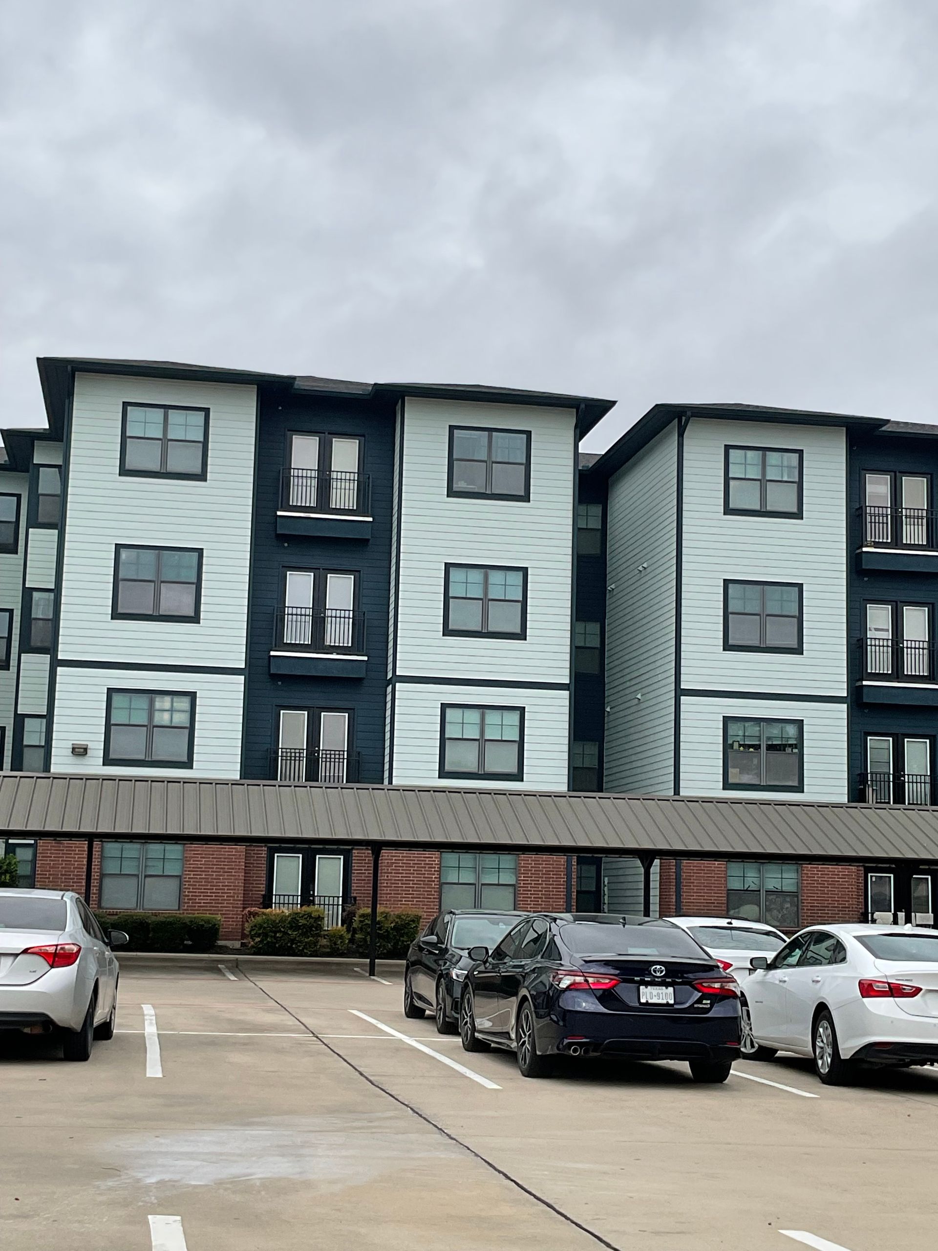 Apartment building with teal and dark blue siding, covered parking, cars parked in front, overcast sky.