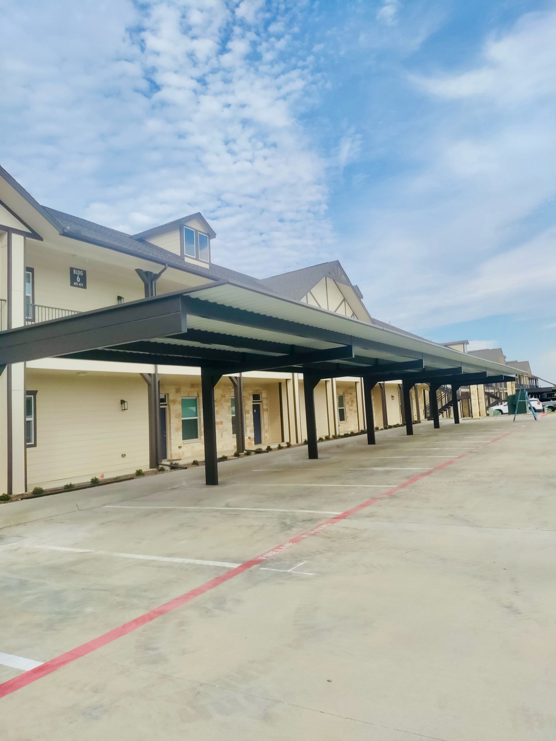 Exterior of a building with a covered parking area. Beige walls, brown canopy, blue sky.