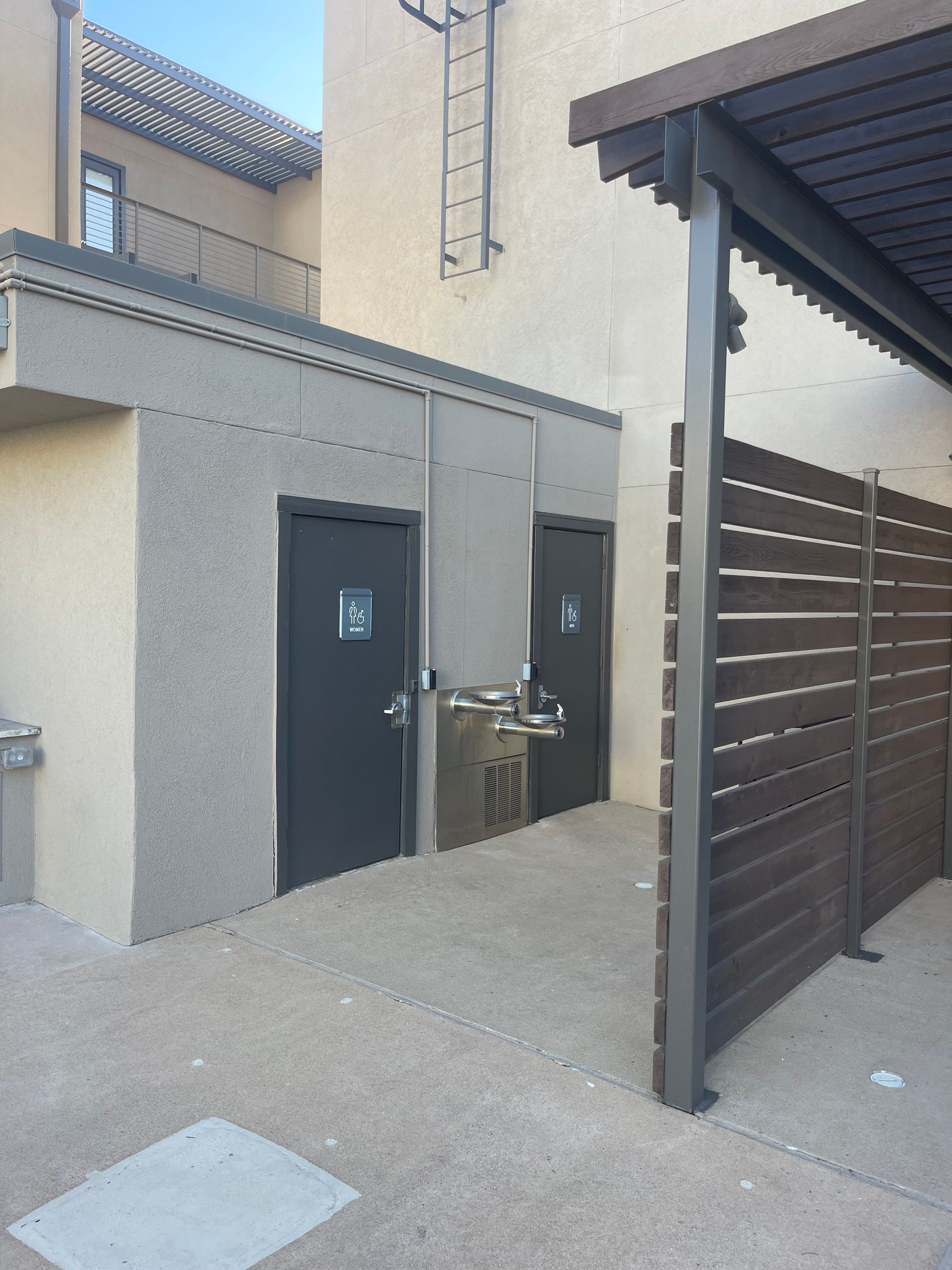 Public restroom building with two doors and a water fountain, next to a shaded area with a wooden fence.