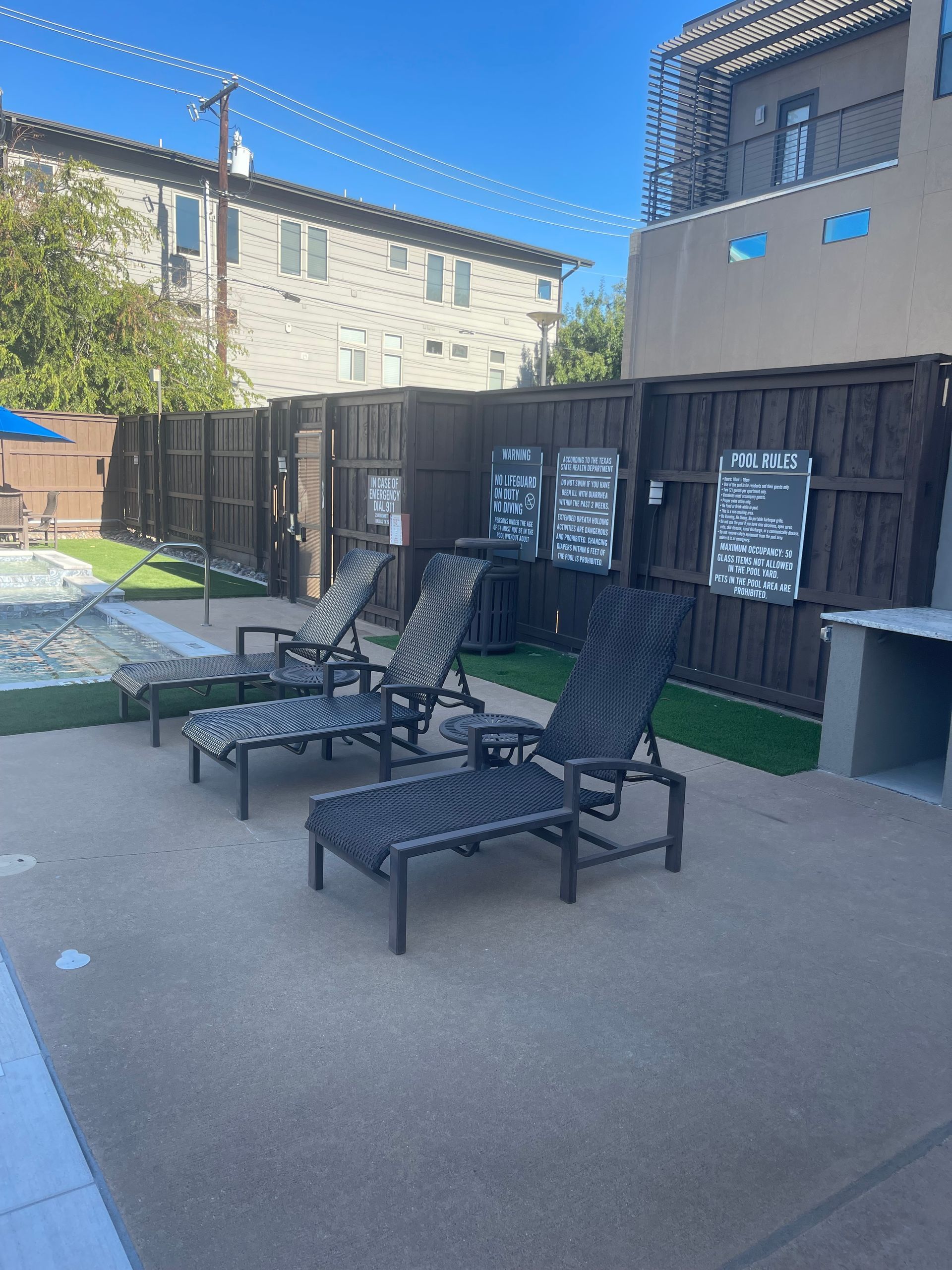 Three black lounge chairs by a pool with a wooden fence background.