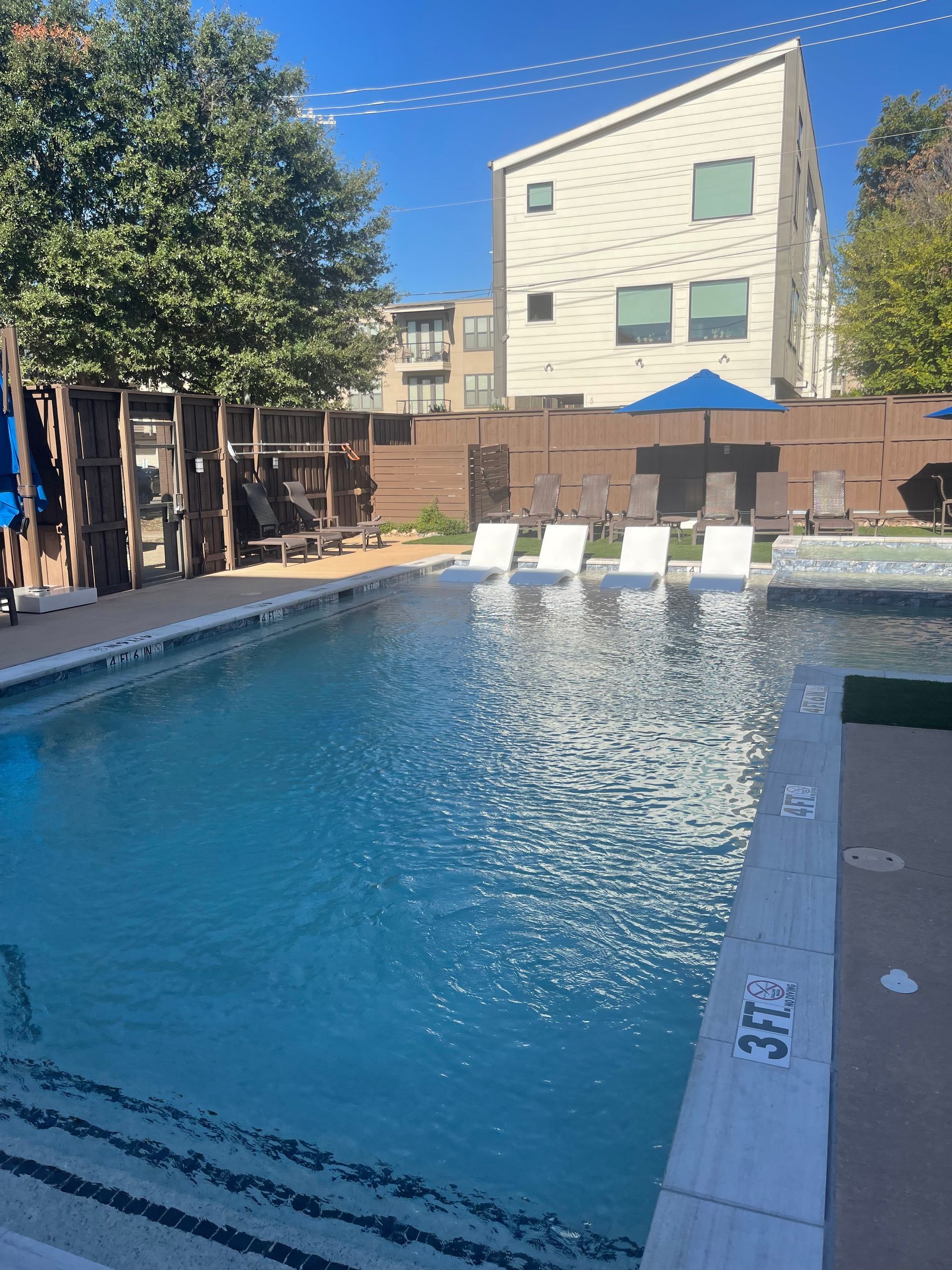 Swimming pool with lounge chairs, blue umbrellas, and a three-story building in the background. Sunny day.