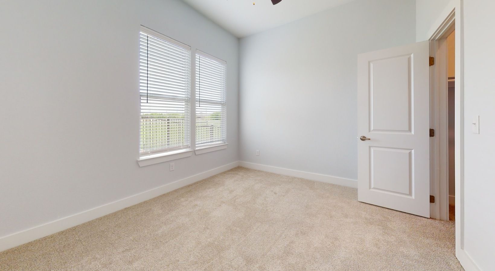 Empty bedroom with a window and a closed door; light-colored walls and carpet.