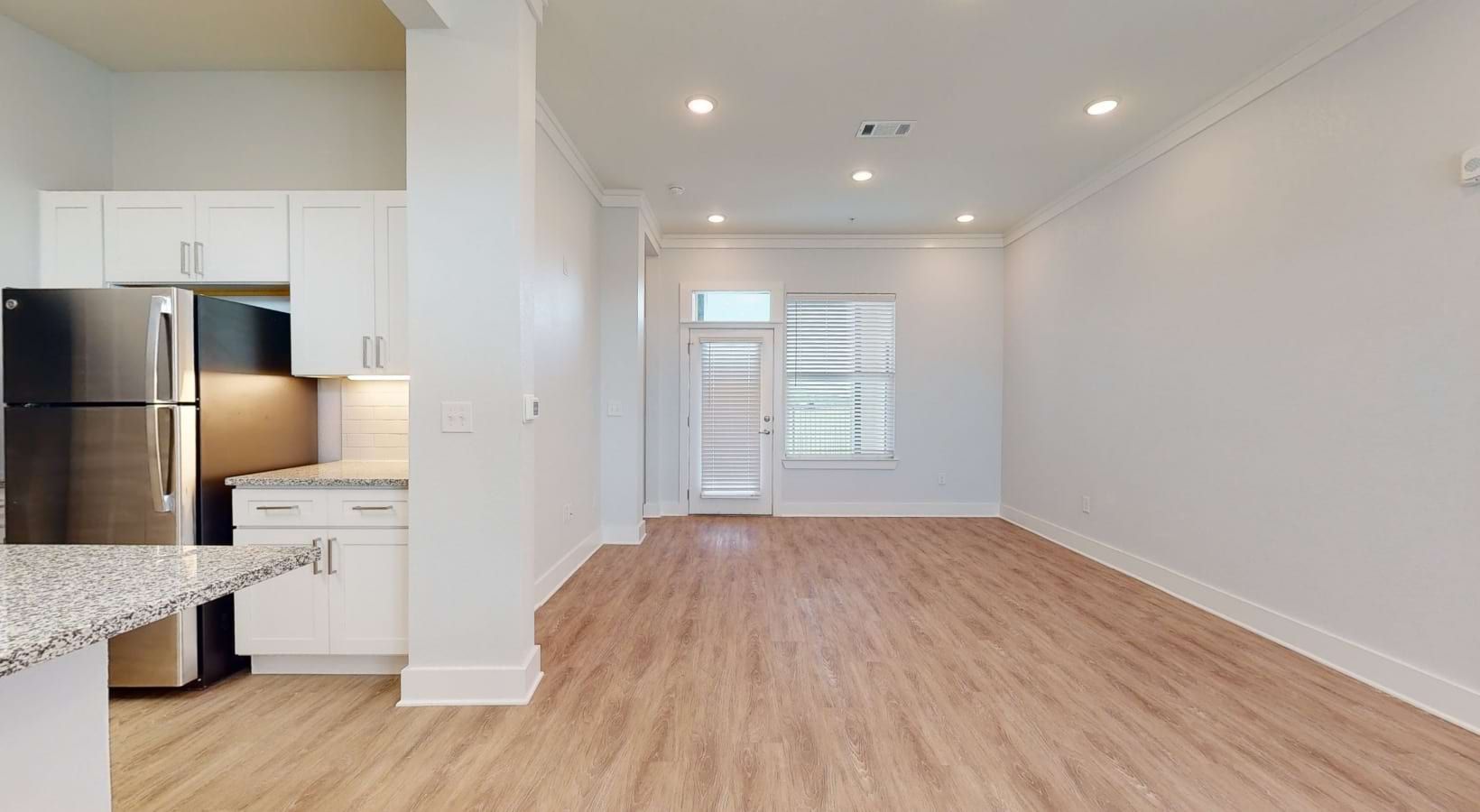 Empty, bright apartment interior: kitchen with white cabinets, stainless steel fridge, light wood floors, and entrance with a door and window.