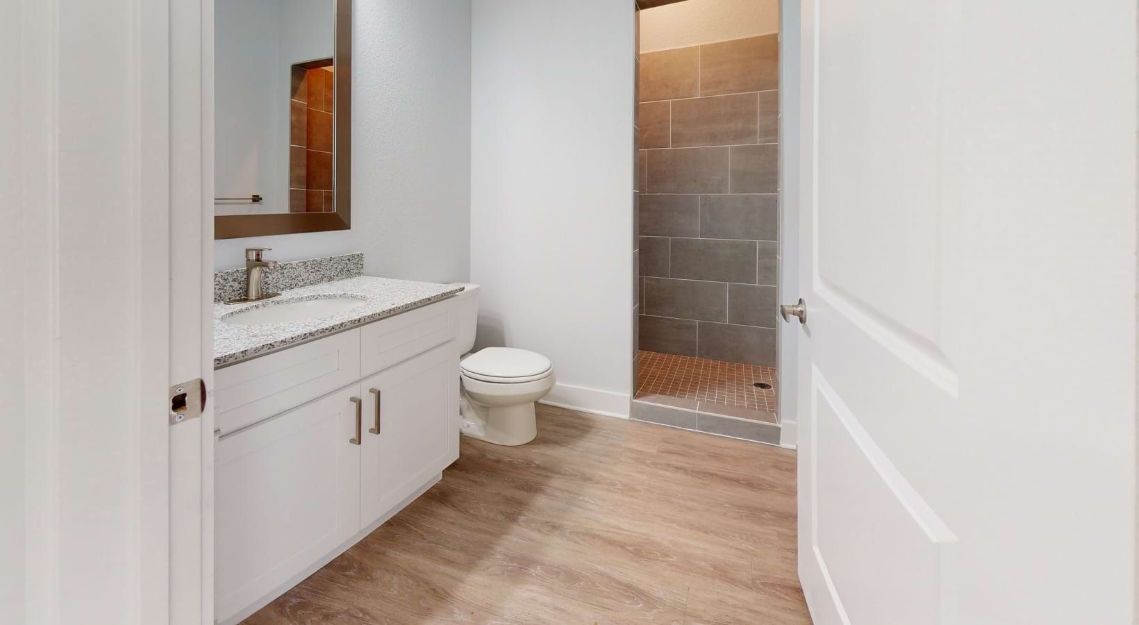 Bathroom interior with a white vanity, toilet, and walk-in shower. Light wood-look flooring and gray shower tiles.