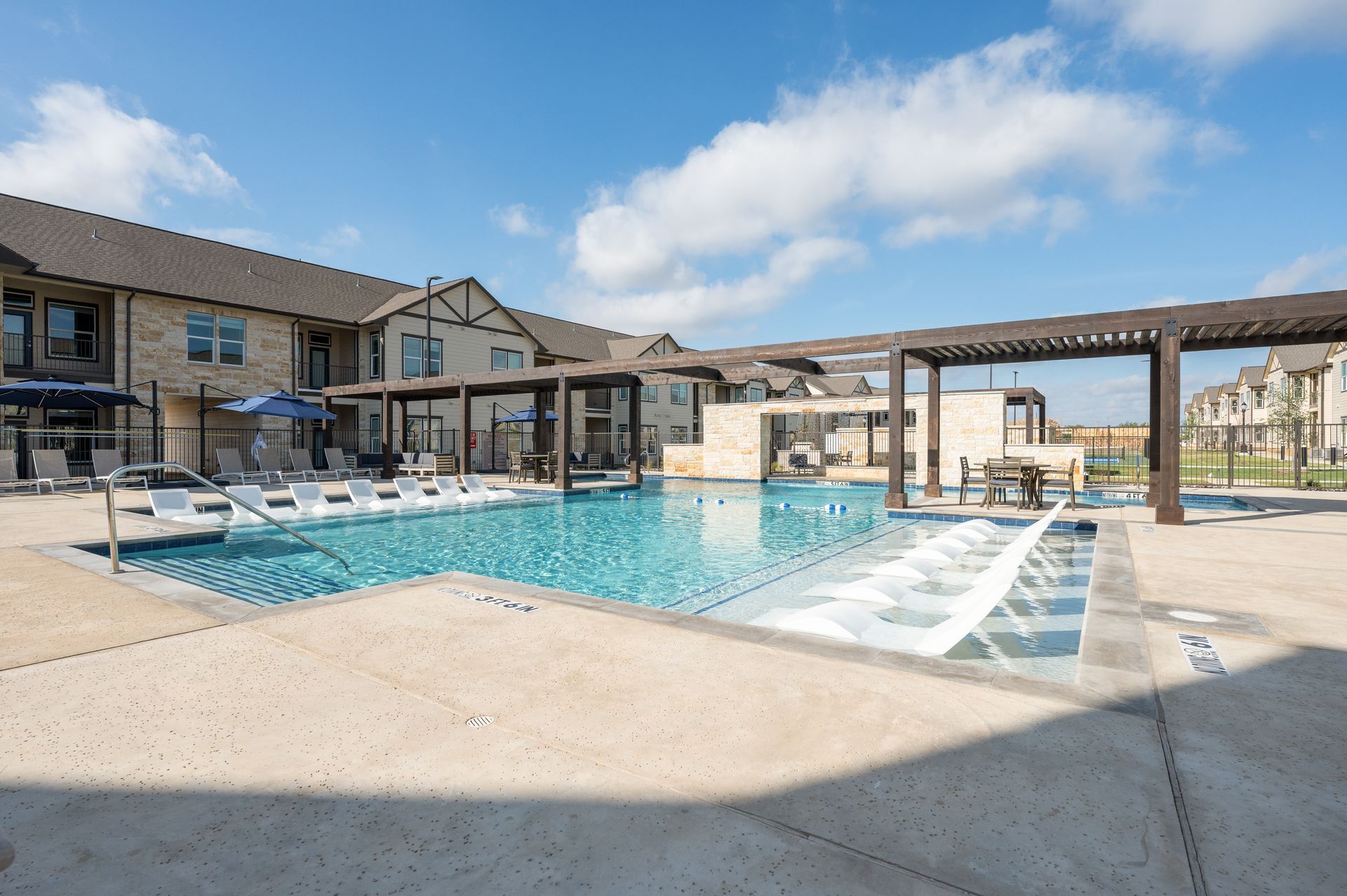 Swimming pool with lounge chairs, pergolas, and a building under a bright blue sky.