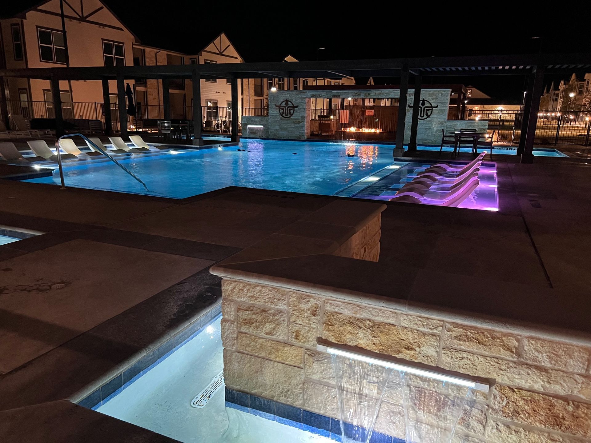 Pool lit up at night with lounge chairs, a bar area, and buildings in the background.