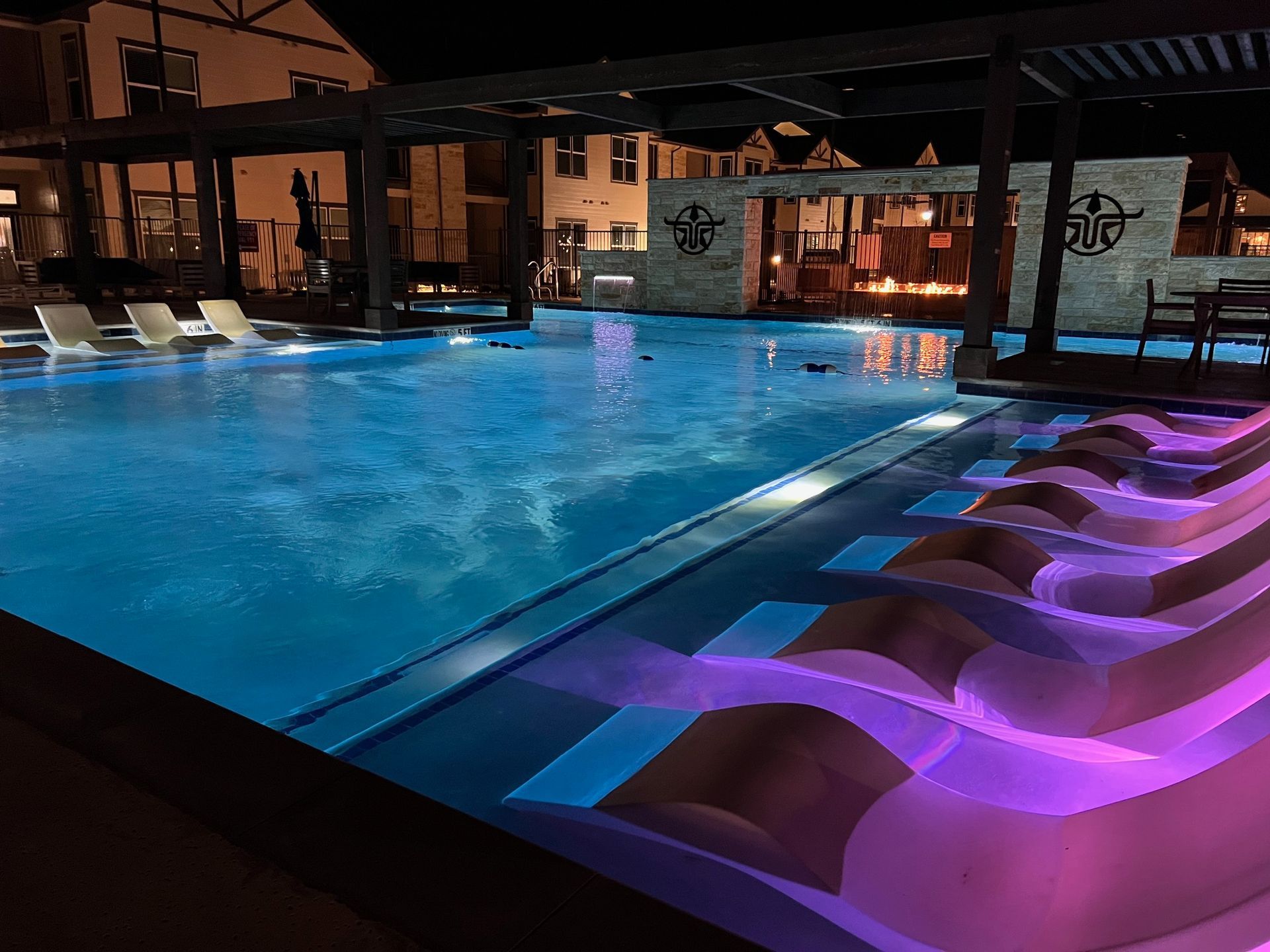 Night view of a lit pool with lounge chairs and a pergola, buildings in the background.