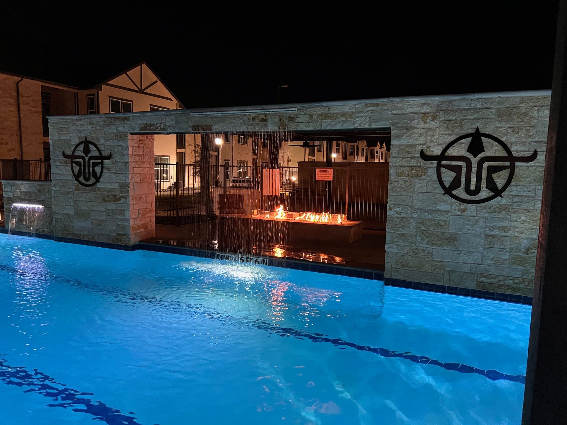 Pool with waterfall feature at night, lit with blue and orange lights, building in background.