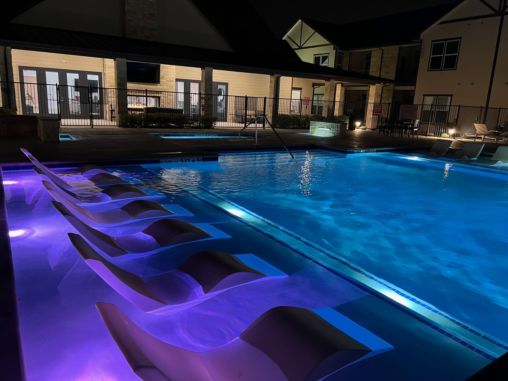 Night view of a lit swimming pool with submerged lounge chairs, next to an open-air building.