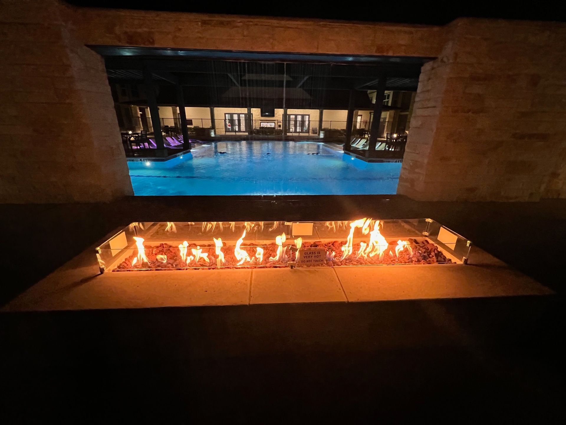 A rectangular fire pit with flames, viewed through a stone opening, and a swimming pool in the background at night.