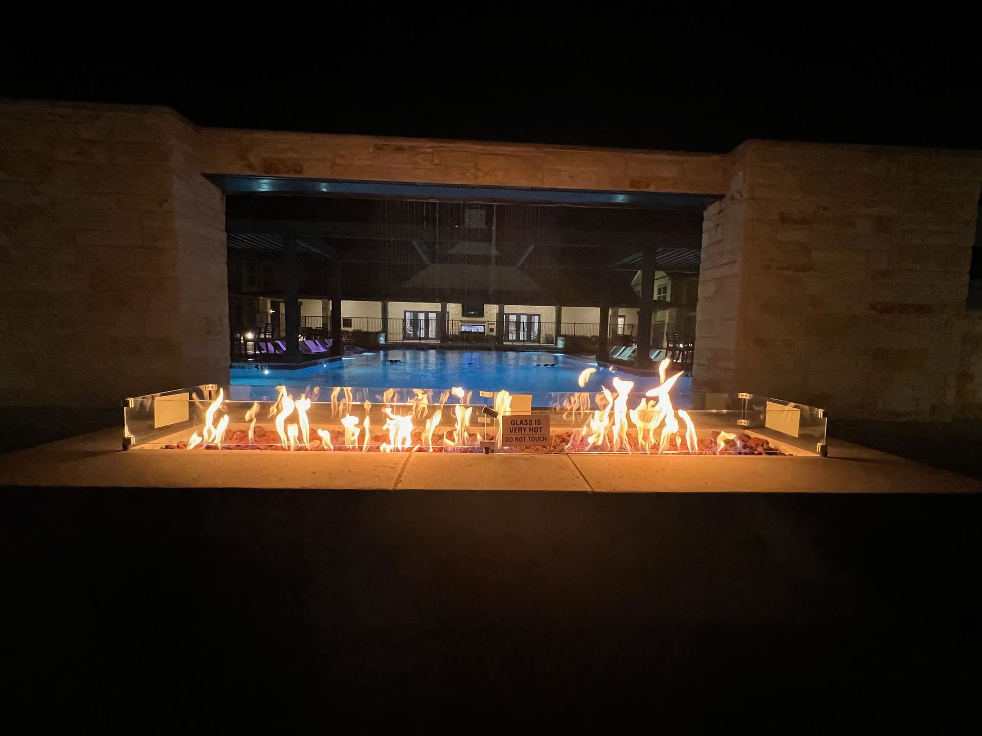 A row of flames burns in front of a pool, viewed through an opening in a stone structure at night.