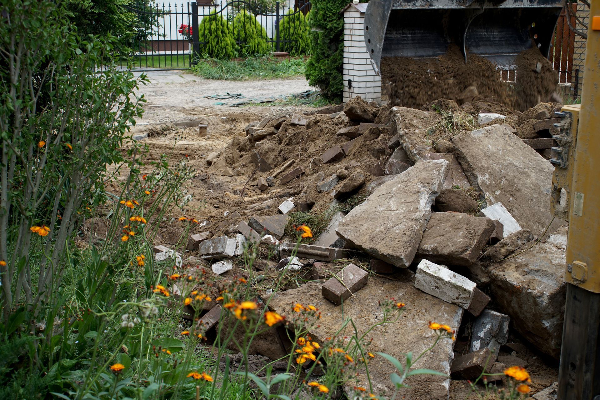 Demolition debris: concrete blocks and dirt pile near a construction site. Green plants and flowers in the foreground.