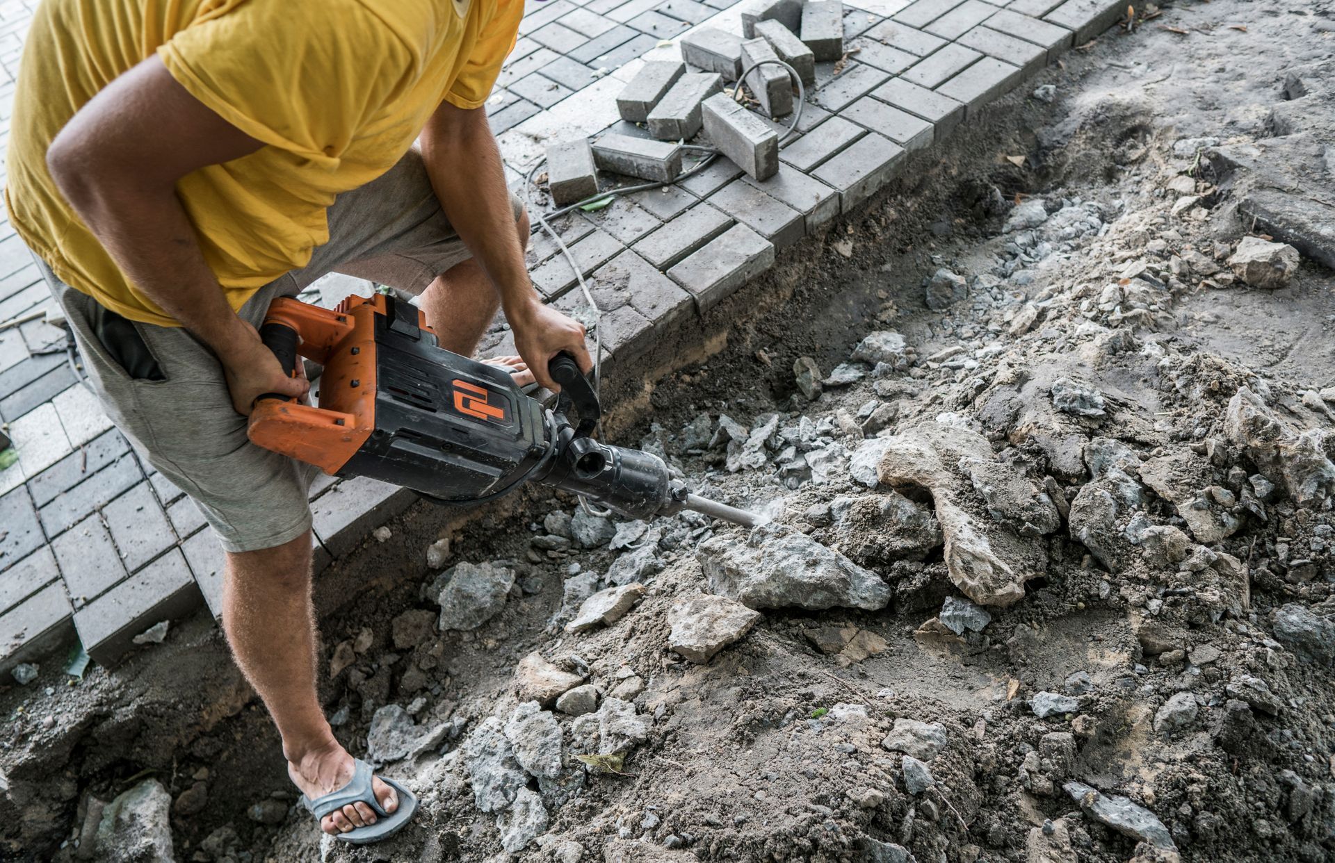 Person using a jackhammer to break up concrete next to a paved area.