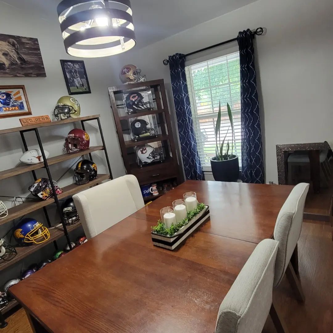 Dining room with wooden table and chairs, shelves of helmets, and window with blue curtains.