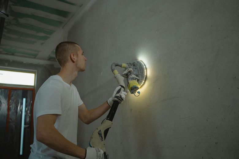 Man sanding a wall with a disc sander, wearing white shirt and gloves in a construction setting.