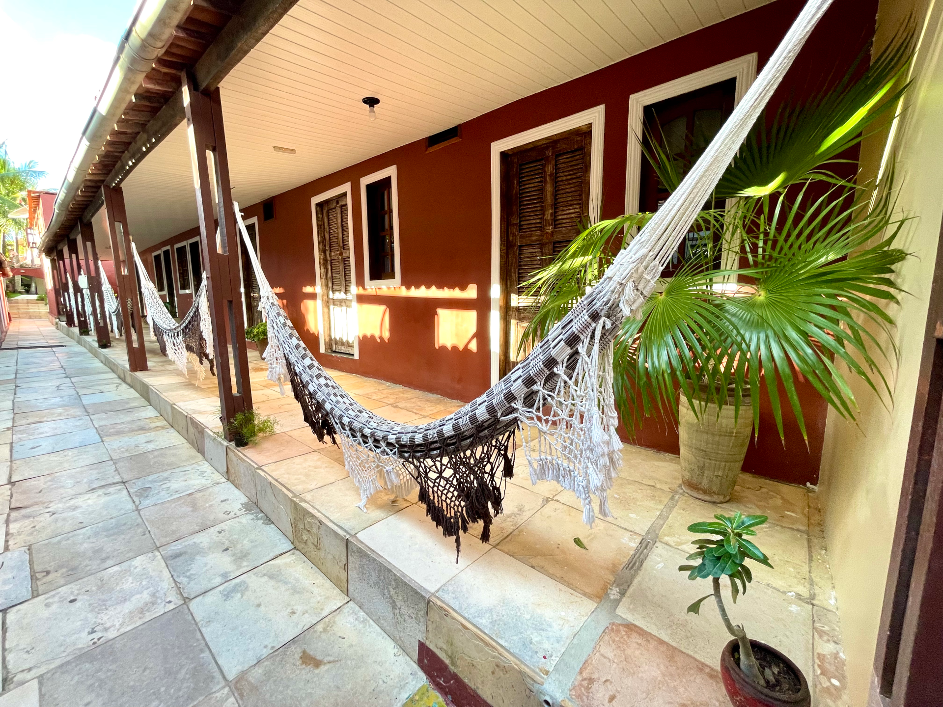 Varanda com redes estendidas entre postes de madeira. Parede vermelha com janelas, vasos de plantas e piso de pedra.