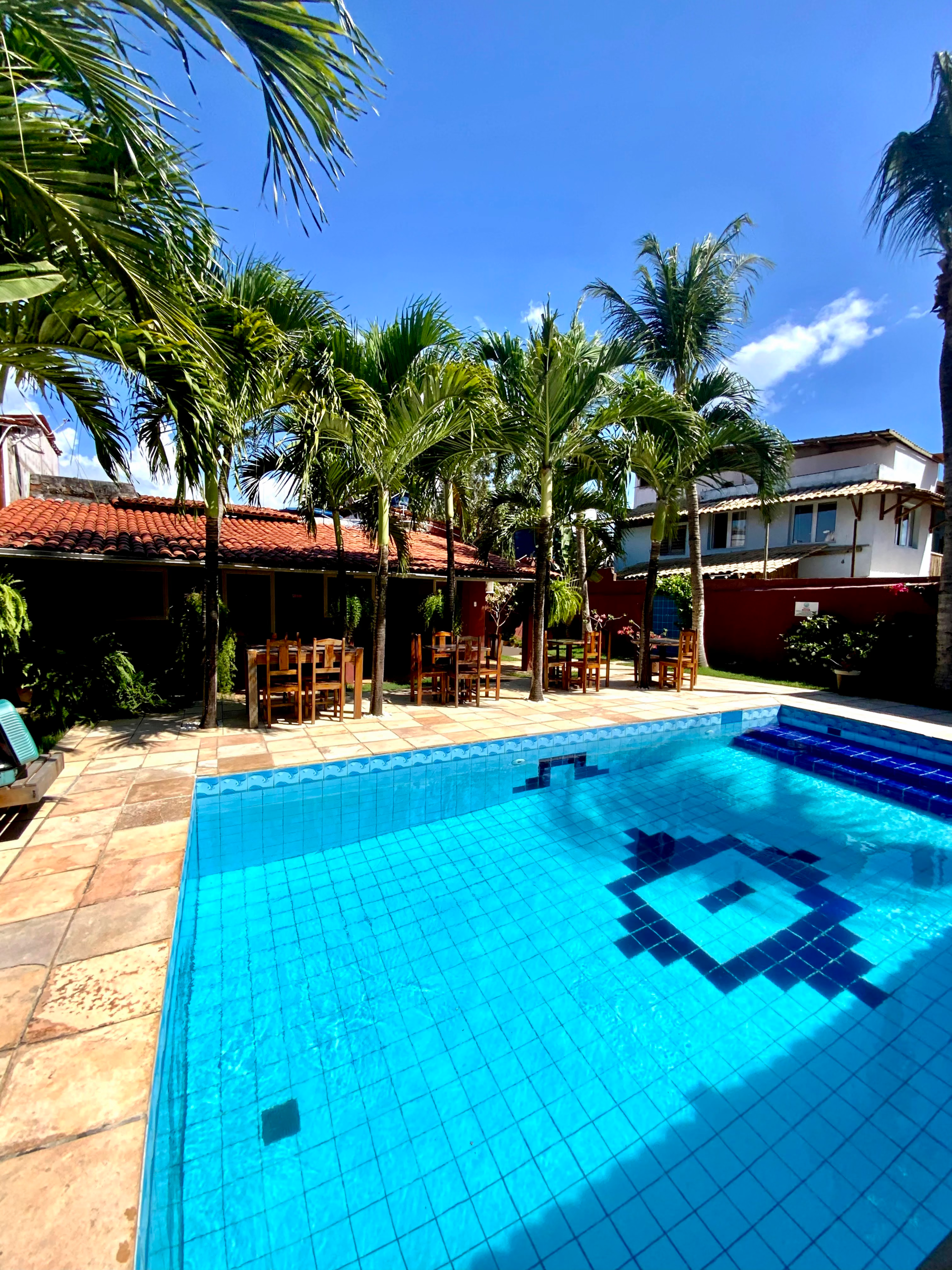 Piscina com água azul, mesas e cadeiras sob as árvores, dia ensolarado.