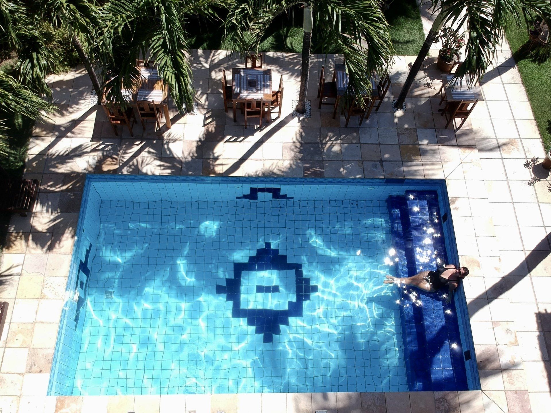 Vista aérea de uma piscina retangular com uma pessoa relaxando. Palmeiras sombreiam mesas e cadeiras próximas.