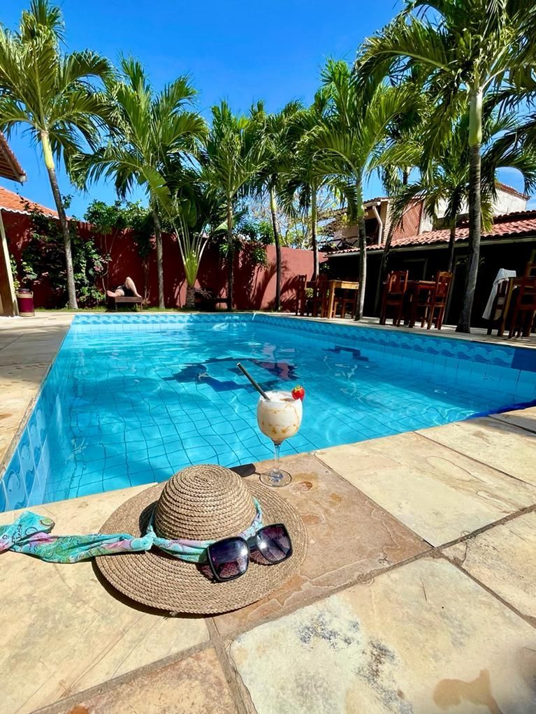Cena à beira da piscina: chapéu, óculos de sol, bebida, piscina azul, palmeiras, dia ensolarado.