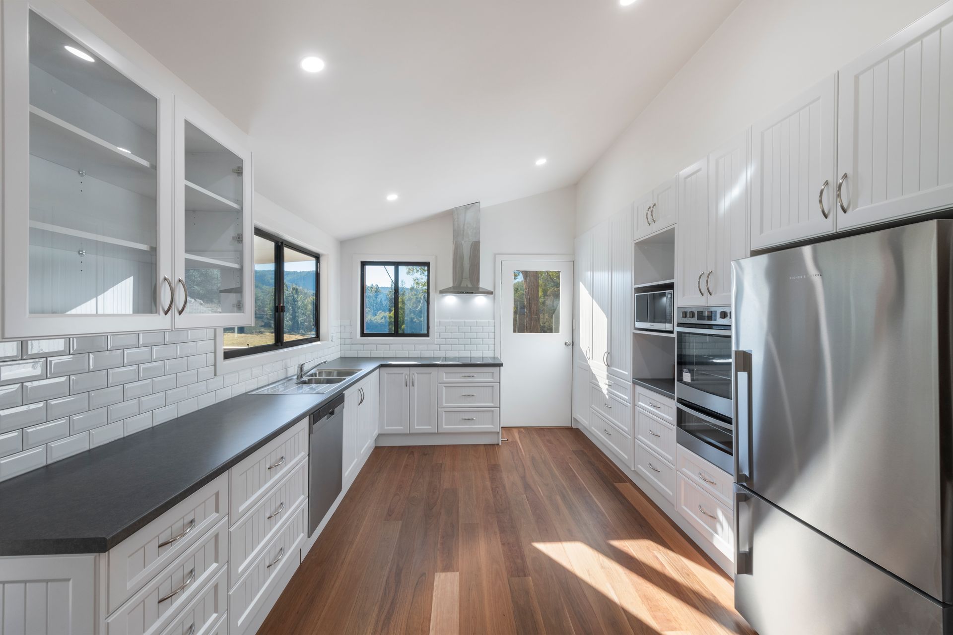 White Kitchen With Wooden Floor, Dark Countertop, and Glass-door Cabinets — Indie Living In Lake Macquarie, NSW
