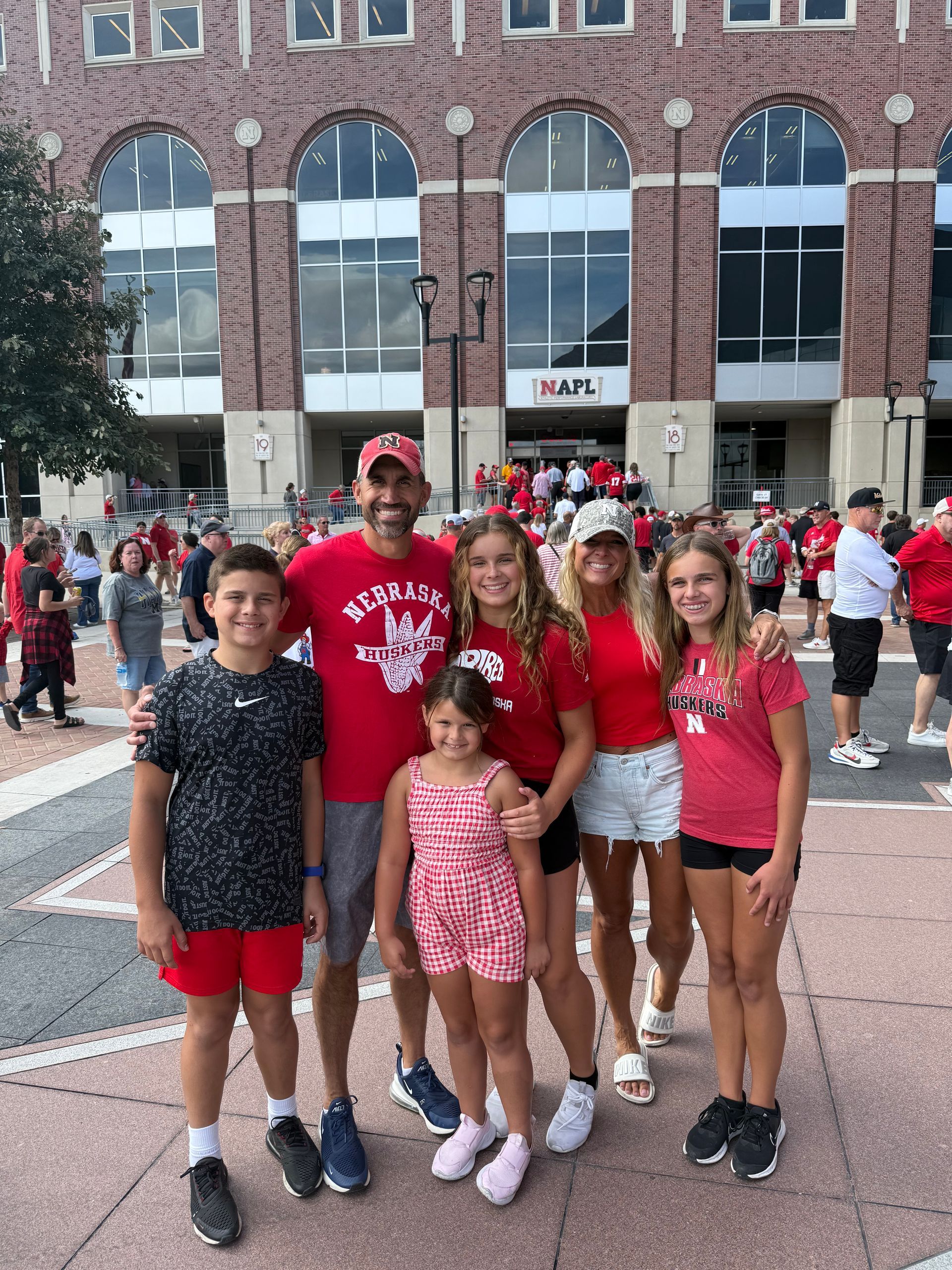 A family poses for a photo outside a large brick stadium, wearing casual clothing and smiling on a sunny day.