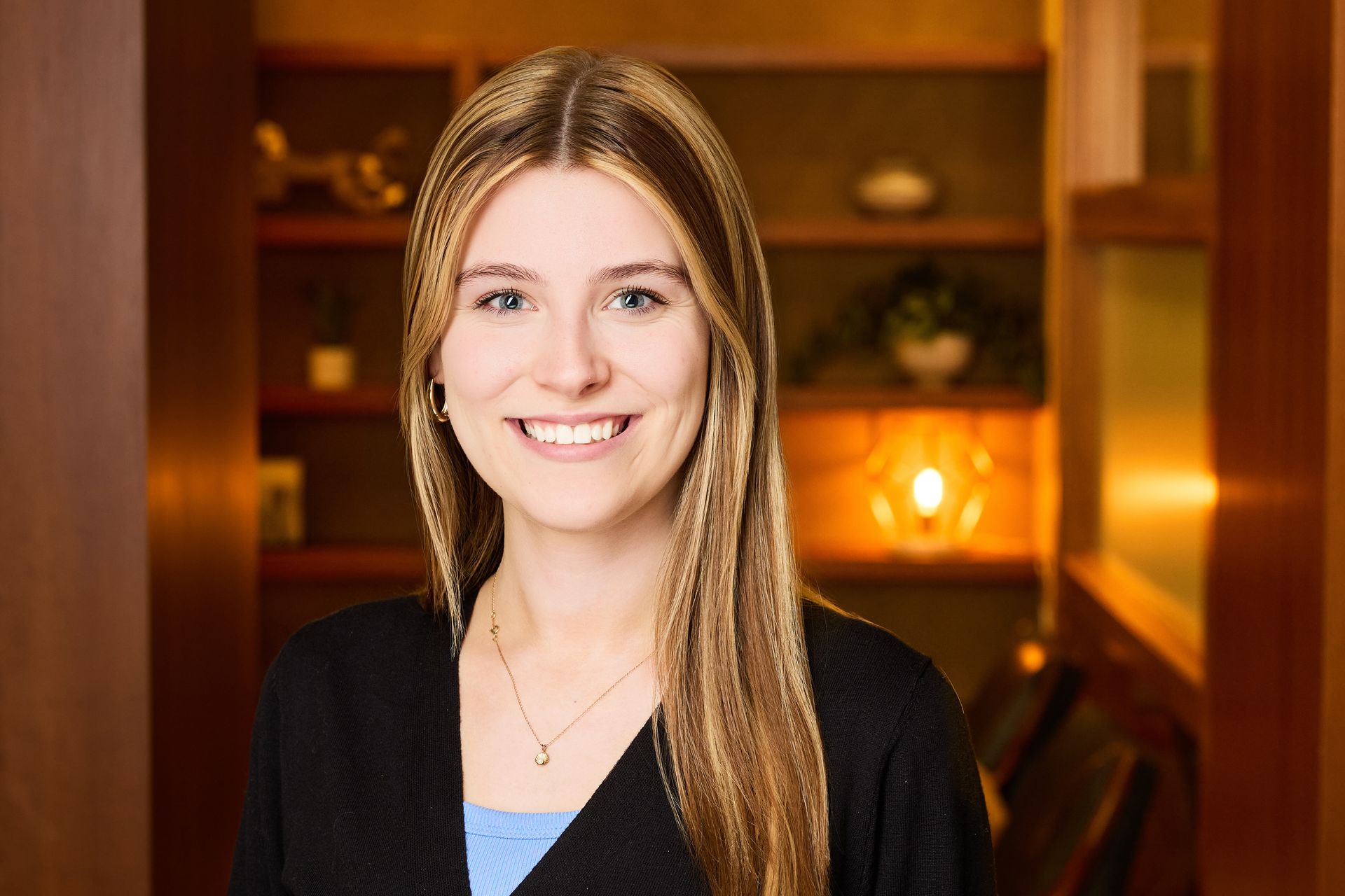 Woman with long brown hair smiles, wearing a black blazer and teal top in an office setting.