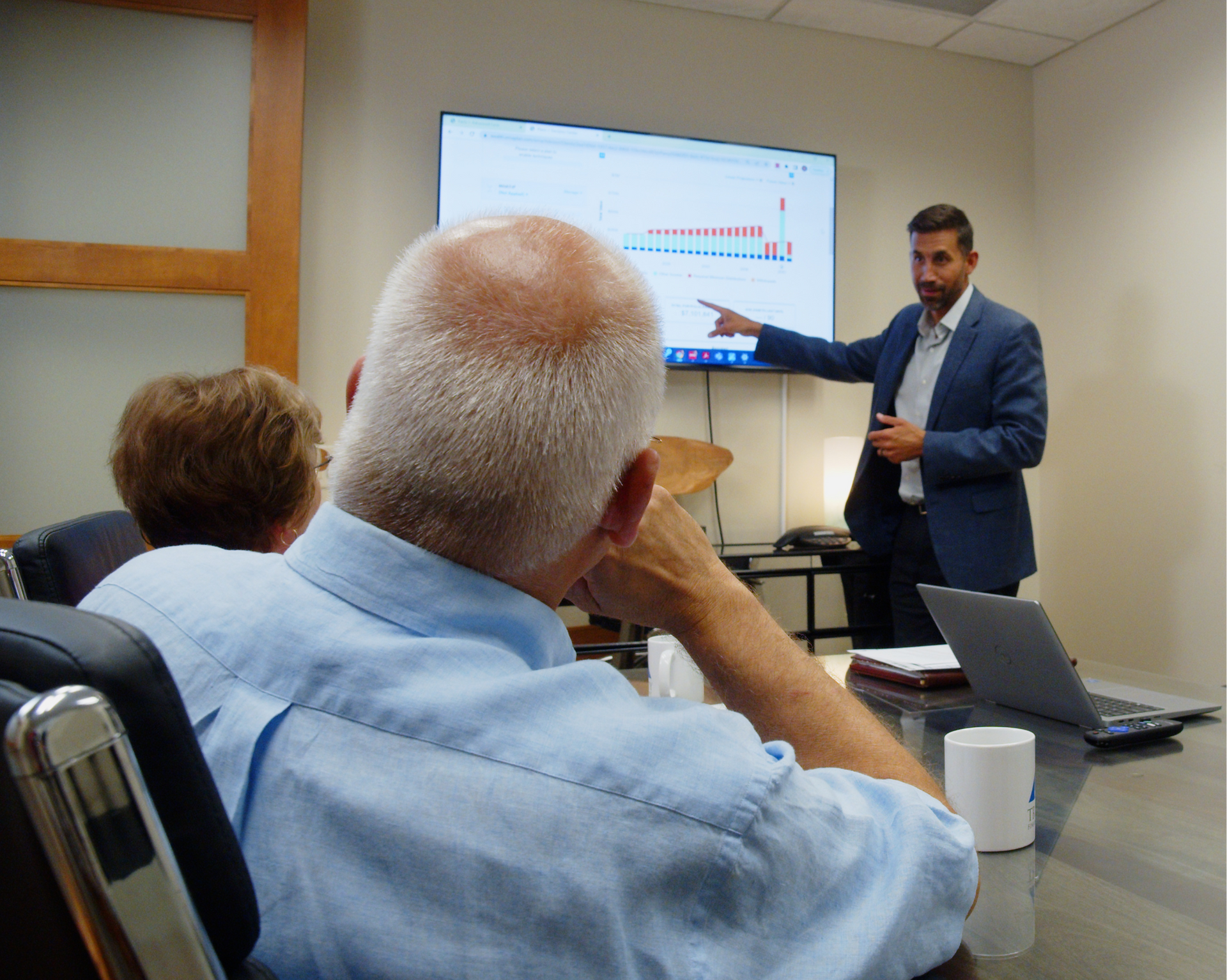A man in a blue suit points at a chart on a wall-mounted screen while presenting to people at a conference table.