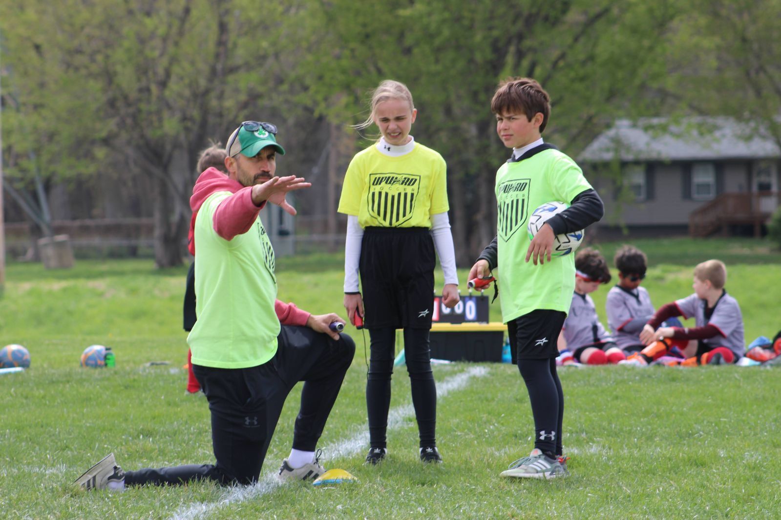A coach in a neon yellow shirt kneels on a field to instruct two players while others sit in the background.