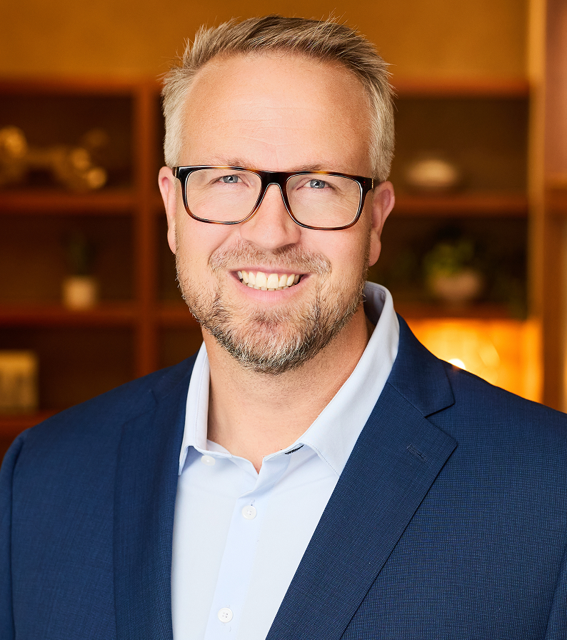 Man in glasses and blue suit smiles in an office setting.