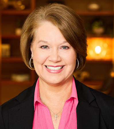 Woman with shoulder-length brown hair, smiling, wearing a pink shirt, black blazer, and cross necklace. Blurred background.