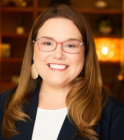 Woman with brown hair and glasses smiling, wearing a blazer, with a blurred background.