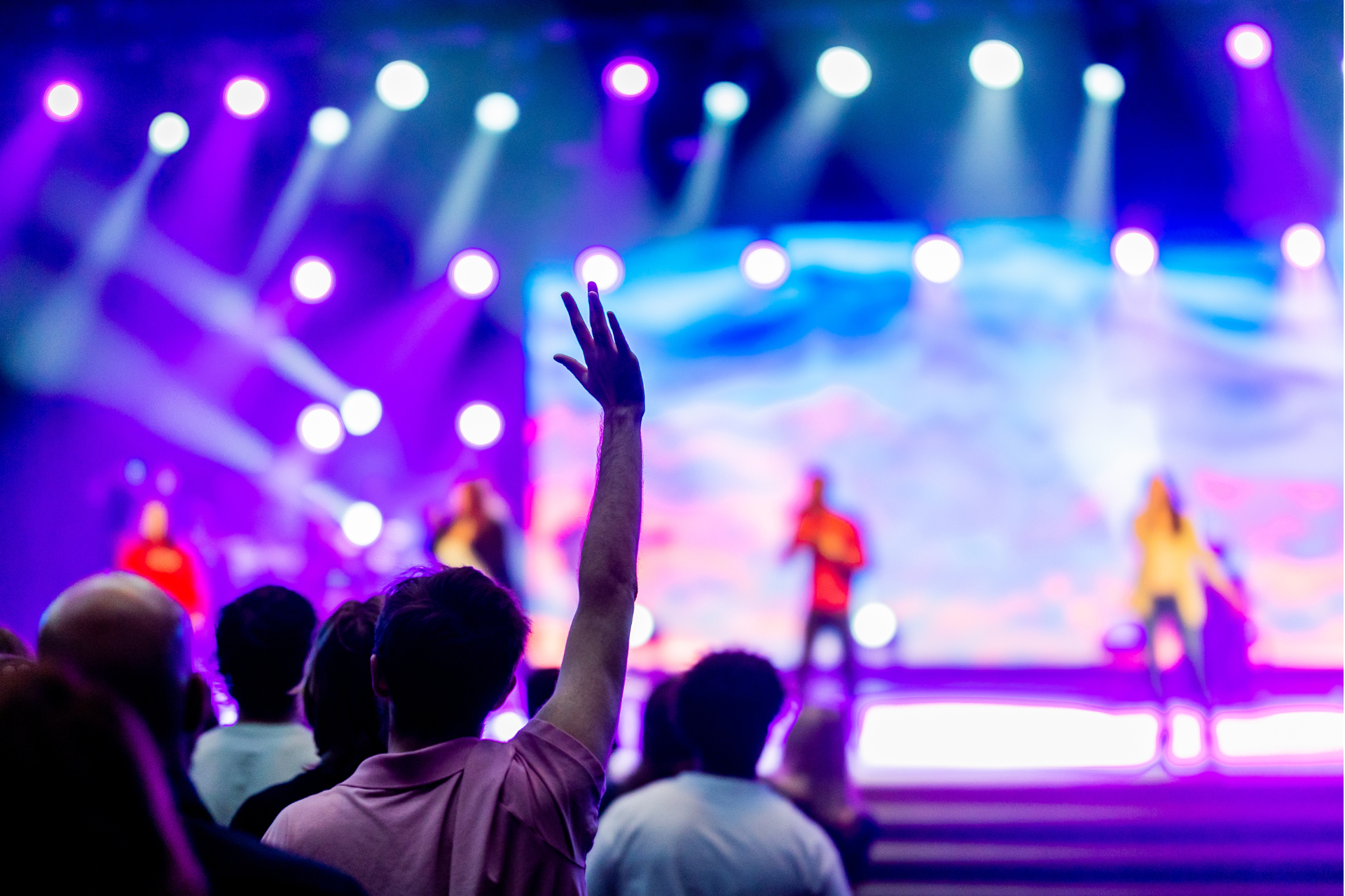 Person with raised arm in a crowd at a concert with performers on stage, under blue and purple lights.