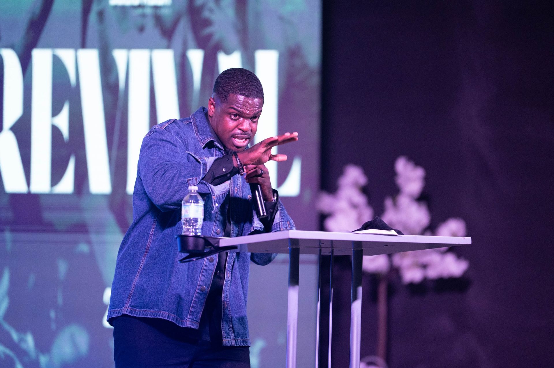 Man in denim jacket speaking at a podium, gesturing with hands. Stage with 