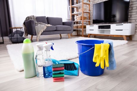 Prodotti per la pulizia appoggiati sul pavimento di un soggiorno moderno, con un secchio blu, un flacone spray e degli stracci.