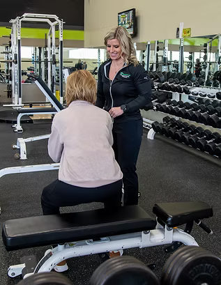 Fitness trainer speaking with a client in a gym beside a workout bench and dumbbells.
