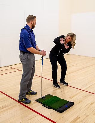 Two people in a gym, one holding a strap while the other bends forward beside a balance board.